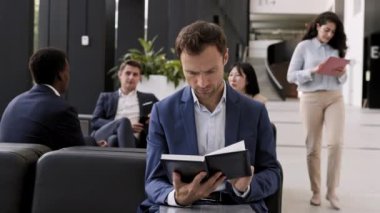 Medium shot of good-looking Caucasian businessman wearing elegant suit sitting on sofa in modern coworking space, looking through notes in notebook and then looking at camera