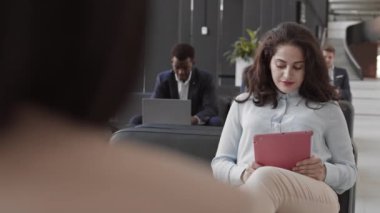 Over the shoulder view of young mixed-race female office worker wearing formalwear sitting on sofa in modern coworking space with digital tablet in hands, looking at camera and smiling