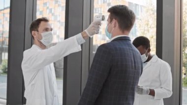 İki meslektaşı sonuçları tablete koyarken, erkek doktorun iş adamlarının iş merkezine kızılötesi termometre kullanarak girmelerinin sıcaklığını kontrol etmesi