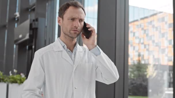中镜头的白种人男性科学家身穿医疗服 沿着现代医院走着 用手机交谈 — 图库视频影像