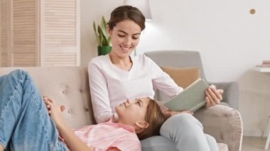 Oturma odasındaki kanepede oturan ve genç kızına başını dizlerinin üzerine çöküp kitap okuyan genç ve çekici bir kadın.