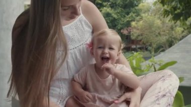 Mavi gözlü, açık havada sarı saçlı, çiçekli, beyaz bir kız çocuğu tutan uzun saçlı genç bir kadının orta boy yakın çekimi.