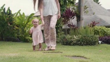 Long shot of cheerful Caucasian baby girl wearing pink dress and fresh flower in hair smiling, walking on green grass in garden, then looking on camera and hugging her long-haired mom