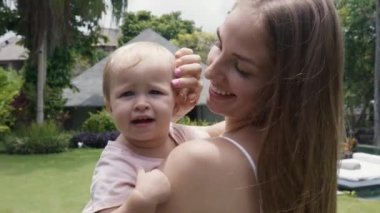 Medium close-up arc POV of long-haired young Caucasian mother holding joyful blue-eyed female toddler in her arms in greenery. Baby daughter smiling, hugging mommy, looking on camera