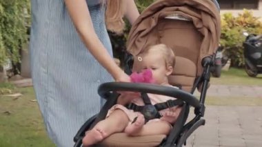 Blond-haired Caucasian one-year-old child sitting in stroller, looking around, unrecognizable mother in striped white and blue dress giving her baby bright pink fresh flower