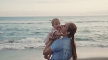 Arc shot of long-haired Caucasian young mother smiling, lifting blue-eyed cute toddler up above her, then hugging, kissing child by seashore