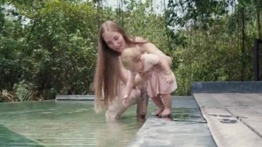 Long shot of beautiful Caucasian mother smiling, sitting ankle-deep in water in artificial pond in courtyard with her toddler daughter, baby girl playing with fallen leaf