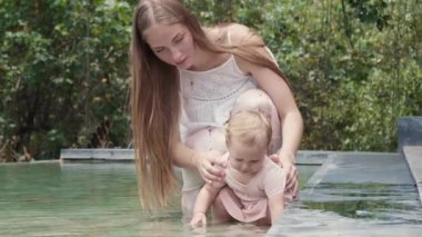 Full shot of long-haired Caucasian young mother smiling, sitting ankle-deep in water in artificial pond in courtyard with her one-year-old daughter, toddler playing with yellow fallen leaf