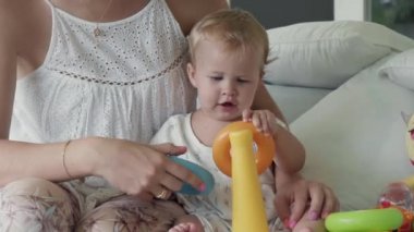 Medium shot of cute Caucasian baby girl playing with colorful toy ring pyramid, sitting with cropped unrecognizable mother