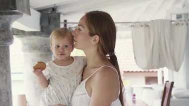 Medium close-up of happy young Caucasian mother walking around terrace, smiling, talking, dancing, holding blue-eyed baby girl eating cookie, then kissing her on cheek