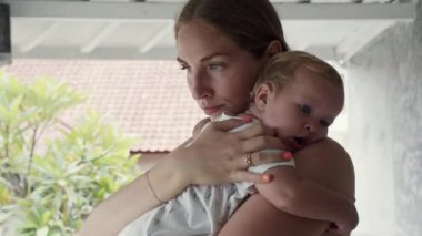 Medium close-up of Caucasian young woman standing on terrace, holding blue-eyed baby girl, toddler yawning, then starting crying out of tiredness