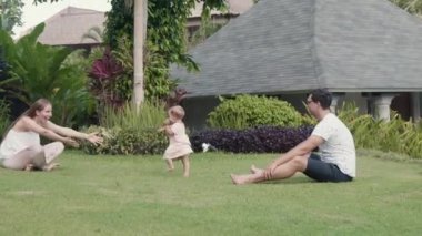 Long shot of two young Caucasian parents sitting on green lawn in garden outside their house, one-year-old child walking from father to smiling long-haired mother, picking baby up