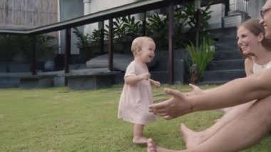 Arc shot of blue-eyed Caucasian toddler in little light-pink dress walking towards young parents sitting on green lawn in garden outside their house. Joyful family smiling, spending time outdoors