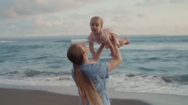 Side view tracking in of long-haired Caucasian young mother smiling, lifting blue-eyed cute toddler up above her, then down, kissing child by ocean coast