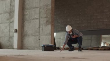 Long shot of professional construction specialist wearing protective goggles and helmet in premises under renovation, holding smartphone in hand, looking at architectural papers on floor