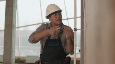 Medium shot of Mixed-Race woman-construction worker wearing helmet and protective goggles, screwing on white plastic pipe with metallic staple to wall