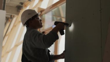 Side view waist-up of young Mixed-Race construction worker wearing helmet and goggles standing indoors, using drill on plasterboard wall, then checking result