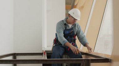 Medium long shot of professional young construction worker wearing helmet and goggles, sitting on metal structure of building under renovation, making measurements with roulette and bubble level