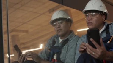 Medium long POV of confident Mixed-Race female construction worker wearing helmet and goggles sitting, leaning on mirror on side, holding smartphone, smiling, her reflection looking up on camera