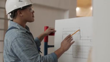 Side view medium close-up of Mixed-Race female professional architect wearing white helmet and protective goggles, standing in construction object, checking by following pencil on blueprint