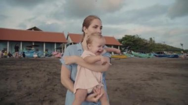 Young Caucasian woman smiling, talking, holding happy toddler, then squatting, pointing finger, showing invisible thing to child. Mother and kid on windy beach