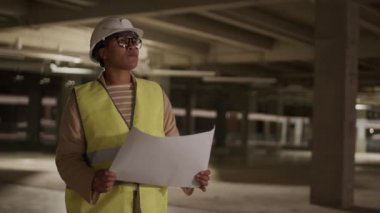 Medium locked-down of Mixed-Race female professional contractor wearing glasses, helmet and vest, holding paper, standing in dark premises under construction, then walking away