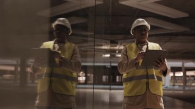 Medium long shot of Mixed-Race construction worker wearing hard hat and glasses holding tablet computer, making notes on it, standing in dark premises in process of renovation near glass wall