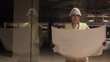 Medium long shot of Mixed-Race construction worker wearing hard hat and glasses holding large piece of paper with blueprint shining through, standing in dark premises in process of renovation