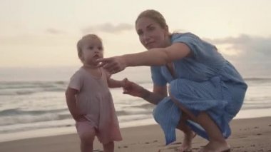 Dollying out of happy blue-eyed Caucasian one-year-old child and long-haired woman in blue dress smiling, walking by seashore on sandy beach at sunset, looking at dogs