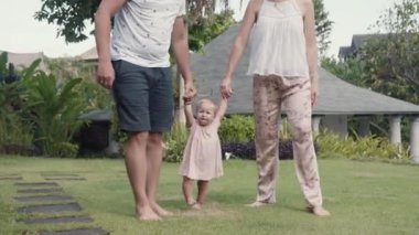 Long shot of two cropped unrecognizable people holding hands with one-year-old Caucasian child in garden, lifting her up. Family walking in greenery