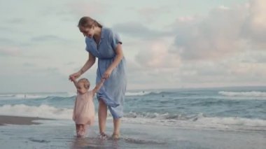 Full shot of happy blue-eyed Caucasian one-year-old baby and long-haired woman in blue dress smiling, walking by seashore on sandy beach, getting wet by waves