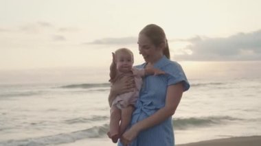 Medium long POV of long-haired Caucasian young mother smiling, holding blue-eyed cute toddler, laughing, joyful child clapping hands on beach by sea at sunset
