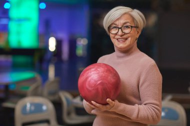 Bowling salonunda eğlenirken elinde bowling topu tutan ve kameraya bakan gülümseyen son sınıf kadınının belden yukarı portresi.