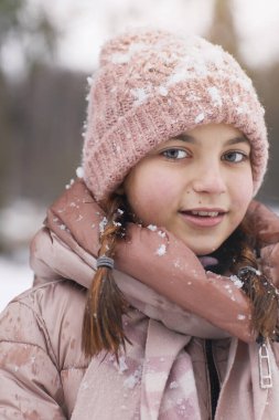 Kışın gülümseyen genç kızın dikey yakın portresi. Pembe şapkalı ve ceketli, karla kaplı kameraya bakarken.