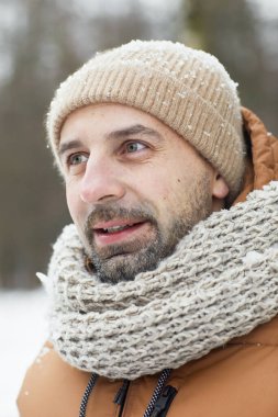 Sakallı olgun bir adamın dikey yakın portresi. Kışın açık havada örgü şapka ve ceket giyip karla kaplı bir şekilde yüzünü çeviriyor.