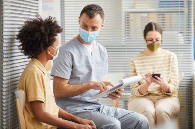 Genç bir doktorun portresi klinikte sırada bekleyen bir çocukla konuşuyor. Maske takıyor, fotokopi çekiyor.