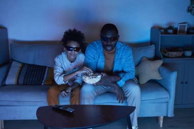 Afrika kökenli Amerikalı bir adam ve genç oğlunun oturma odasında oturup 3 boyutlu film izlediği ve akşam patlamış mısır yediği yüksek açılı bir çekim.