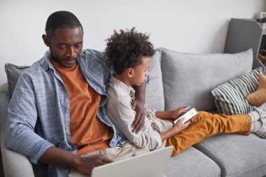 Modern Afrikalı Amerikalı bir adam ve genç oğlunun oturma odasında dizüstü bilgisayar ve akıllı telefon kullanarak rahat bir şekilde oturduğu yüksek açılı bir fotoğraf.