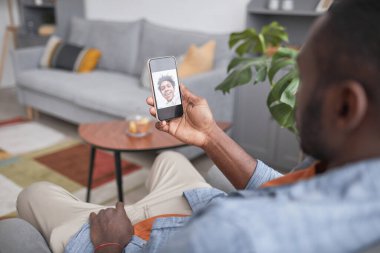 Modern Afrikalı Amerikalı adam kanepesinde oturmuş akıllı telefonuyla neşeli oğluyla görüntülü konuşma yapıyor.