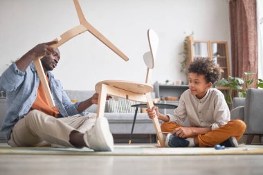 Afrikalı Amerikalı bir aile babası, oğluyla oturma odasında kırık sandalyeyi tamir ederek vakit geçiriyor.