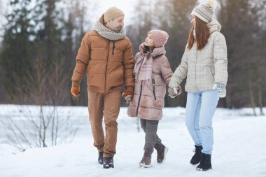 Güzel kış ormanlarında yürürken el ele tutuşup gülümseyen modern mutlu ailenin tam boy portresi.
