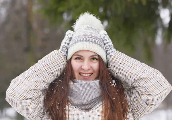 Front view portrait of happy young woman in winter outdoors wearing knit hat and mittens while smiling at camera covered in snow