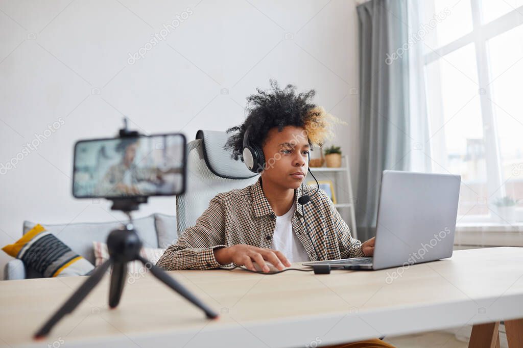 Portrait of teenage African-American boy wearing headset and using laptop while streaming video games at home, young gamer or blogger concept, copy space