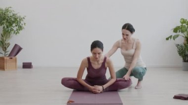 Kadın yoga koçu, kauçuk mindere oturmuş bacaklarında esneme egzersizleri yaparken beyaz kadın sporcuya yardım ediyor.