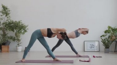 Yoga minderlerinin üzerinde spor giysiler giyen ve içeride yoga yaparken ellerinin arkasında poz veren iki kadın yoginin geniş açılı görüntüsü.