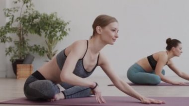 Spor kıyafetleri giyen, spor giyinen, spor salonundaki lastik paspaslarda yan yana oturan ve vücutlarını esneten iki güzel beyaz kadın.