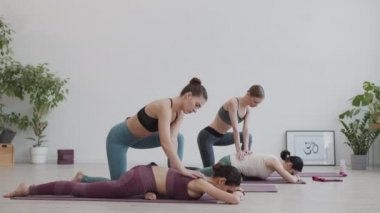Wide shot of two Caucasian attractive sporty women lying in special pose on rubber mats, stretching their backs while two other women pressing on their backs with her hands