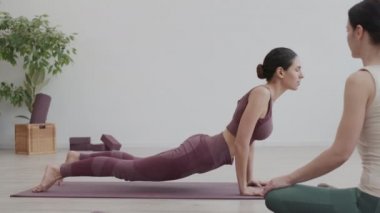 Orta yaşlı kadın koçun yoga minderinde oturup spor giyerek hatha yoga egzersizleri yapan genç bir kadını izlemesi.