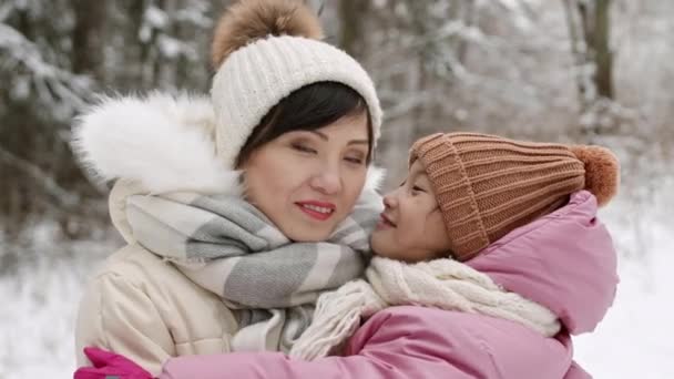 雪の冬の公園でポーズをとっている陽気なアジアの母親と子供の中間クローズアップPov ポンポンやスカーフ付きの帽子 娘さんがママにキスしてカメラを見て — ストック動画