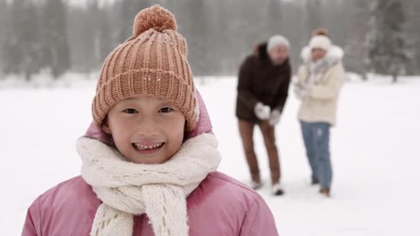 中近接Povの幸せなアジアの子供を着てニットスカーフと帽子でザーメン カメラを見て ぼやけた認識できない人々が彼女の後ろに立って 彼女の上に雪玉を投げます — ストック動画